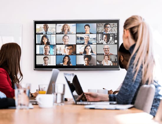 Personeel in een vergaderzaal kijkt naar een scherm met een lopende videoconferentie